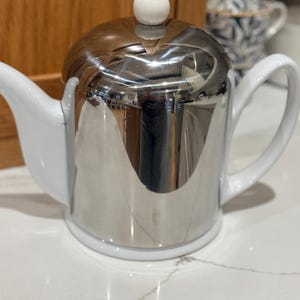 Pode incluir: Um bule com corpo metálico prateado polido e base, pega e bico em cerâmica branca. A tampa é rematada com um pequeno botão redondo de cerâmica branca. A superfície refletora do metal apresenta um reflexo distorcido do ambiente.