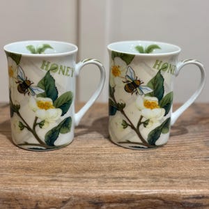 May include: Two white ceramic mugs with a floral and bee design. The mugs feature a cream-colored background with white flowers, green leaves, and a bee. The word "HONEY" is printed on the side of each mug. The mugs are on a wooden surface.