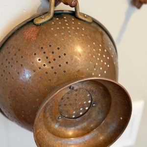 Puede incluir: Un colador de cobre vintage con su base a juego. El colador tiene forma redonda con numerosos agujeros pequeños para escurrir. Cuenta con un asa de latón y está suspendido de un estante de madera. El cobre tiene una pátina cálida y envejecida.