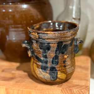 May include: A handmade ceramic jar with a rustic, glazed finish. The jar has a dark brown and black colour scheme with two small handles. The jar is on a wooden chopping board. Another larger jar is in the background.
