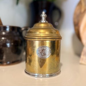 May include: A cylindrical brass canister with a polished silver-toned lid finial and base rim. The front features a circular silver-toned emblem embossed with the text Mermaid Trademark and a graphic of a mermaid figure holding a trident.