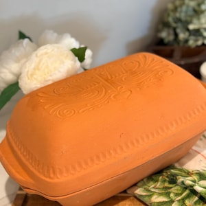 May include: A rectangular terracotta baking dish with a matching lid. The lid features an embossed design of two birds facing each other with floral motifs and a heart in the center, surrounded by a decorative scalloped border along the edges.