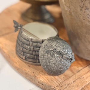 May include: A decorative silver-toned basket-shaped container with a lid, featuring a bee design. The container sits on a wooden tray, with a pedestal bowl and a large vase in the background. The container is open, revealing a white interior.