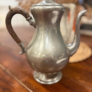 May include: A vintage silver-toned metal coffee pot with a wooden handle and lid finial. The pot has a curved spout and sits on a small pedestal base. The handle is made of dark wood. The pot is likely used for serving coffee or tea.