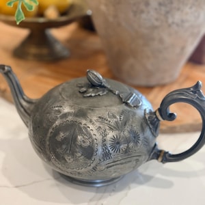 Op de afbeelding: Antieke zilveren theepot met ingewikkelde bloemen- en bladerpatronen. De theepot heeft een gebogen tuit, een ronde body en een handvat met gouden accenten. Het deksel heeft een decoratieve knop. Een klassieker voor theeliefhebbers.
