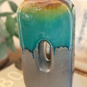 May include: A rectangular ceramic vase with a rounded top and a central vertical cutout. The upper portion features a glossy teal and brown drip glaze, while the lower section has a textured, matte metallic gray finish with a smooth transition.