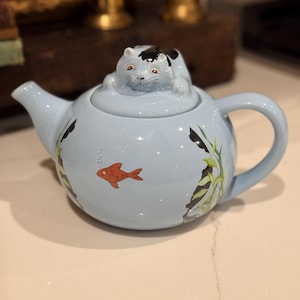 以下が含まれることがあります： 猫の形をした蓋が付いた、水色のセラミックティーポット。ティーポットには、赤い金魚と水生植物のデザインが施されています。ハンドルと注ぎ口も水色です。装飾的なキッチンアイテムです。