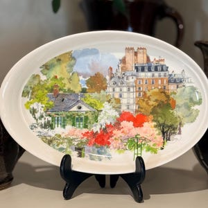 May include: Oval serving platter with a watercolour painting of a European cityscape. The artwork features a house, buildings, and lush greenery in shades of green, red, and orange. The platter is white with a black stand.