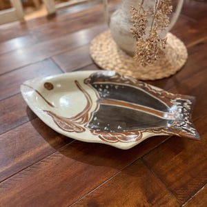 May include: A ceramic fish-shaped serving dish with a beige base and brown and black painted details. The dish has a wide, shallow design and is placed on a wooden surface. The fish has a detailed fin and eye.