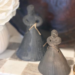 May include: Two dark gray, cone-shaped clay figurines with rounded heads and arms crossed over their chests. Each figure features a thin, light-colored string tied around its waist, creating a simple, textured appearance with a matte finish.