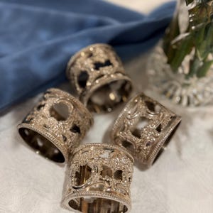May include: Four silver napkin rings with ornate, openwork designs. The rings are arranged on a white tablecloth with a blue cloth in the background. A crystal vase with greenery is visible in the background.