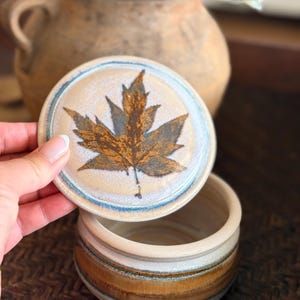 Könnte beinhalten: Ein Keramiktopf mit Deckel, verziert mit einem braunen Ahornblatt. Der Topf hat ein beige-braunes Streifenmuster. Der Deckel wird angehoben und gibt den Blick ins Innere frei. Der Topf ist ein dekorativer Gegenstand.