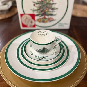以下が含まれることがあります： 白と緑のクリスマスをテーマにした食器セット。カップ、ソーサー、緑の縁取りのあるお皿が含まれています。金色のチャージャープレートの上にセットされています。背景にはクリスマスツリーのイラストが描かれた箱があります。