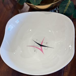 May include: A white ceramic bowl with a slightly irregular, rounded square shape. The interior features a centered graphic design consisting of a black starburst-like shape layered over a pink V-shaped element, accented with small black dots and swirls.