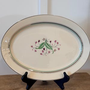 May include: Oval serving platter with a cream-colored surface, a silver rim, and a green and black border. The center features a floral design with pink and green accents. The platter is displayed on a black stand.
