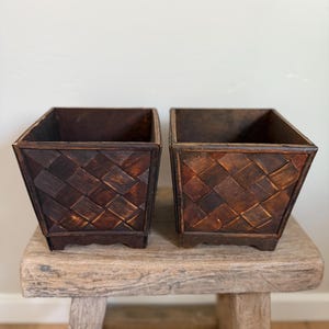 May include: Two square, dark brown wooden planters with a woven pattern. The planters are set on a weathered, light brown wooden stool. The planters have a rustic, natural aesthetic, suitable for indoor or outdoor use.