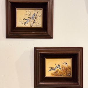May include: Two framed paintings of birds in flight. The paintings feature a warm, yellow background with brown and blue birds. The frames are dark brown and rectangular. One painting shows birds near bare branches, the other shows birds near autumn foliage.