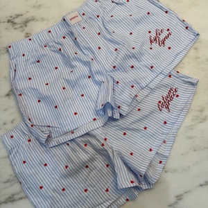 Puede incluir: Dos pares de pantalones cortos de pijama a rayas azules y blancas con detalles de corazones rojos. Un par tiene el nombre "kylie lynn" bordado en rojo, y el otro "tatum grace". Los pantalones cortos tienen cintura elástica.