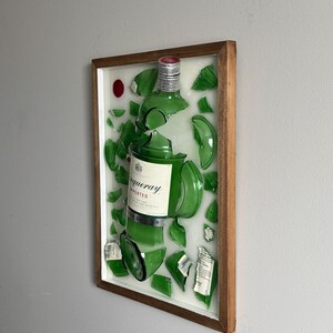 K&ouml;nnte beinhalten: Ein gerahmtes Kunstwerk mit einer zerbrochenen gr&uuml;nen Glasflasche, deren Fragmente auf wei&szlig;em Hintergrund angeordnet sind. Das Etikett der Flasche lautet "Tanqueray Imported". Der Holzrahmen umgibt das Kunstwerk, und ein roter Punkt ist oben links sichtbar.