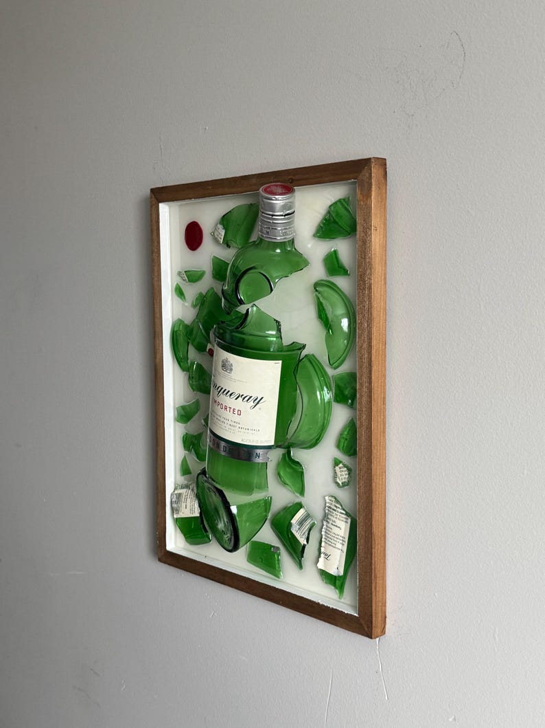 K&ouml;nnte beinhalten: Ein gerahmtes Kunstwerk mit einer zerbrochenen gr&uuml;nen Glasflasche, deren Fragmente auf wei&szlig;em Hintergrund angeordnet sind. Das Etikett der Flasche lautet "Tanqueray Imported". Der Holzrahmen umgibt das Kunstwerk, und ein roter Punkt ist oben links sichtbar.