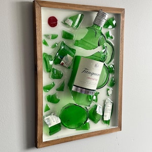 K&ouml;nnte beinhalten: Gerahmtes Kunstwerk mit einer zerbrochenen gr&uuml;nen Glasflasche Tanqueray London Dry Gin. Die Glasscherben sind auf wei&szlig;em Grund in einem Holzrahmen angeordnet. Ein rotes rundes Element ist ebenfalls vorhanden.