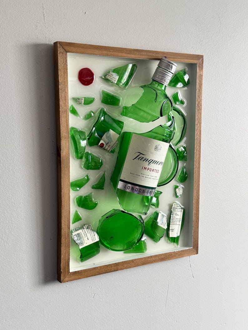 K&ouml;nnte beinhalten: Gerahmtes Kunstwerk mit einer zerbrochenen gr&uuml;nen Glasflasche Tanqueray London Dry Gin. Die Glasscherben sind auf wei&szlig;em Grund in einem Holzrahmen angeordnet. Ein rotes rundes Element ist ebenfalls vorhanden.