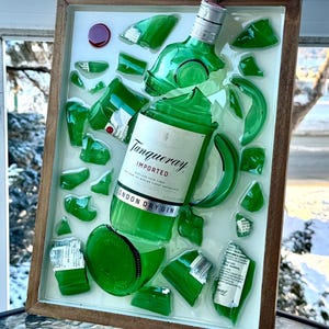 K&ouml;nnte beinhalten: Ein gerahmtes Kunstwerk mit einer zerbrochenen gr&uuml;nen Gin-Flasche, auf der das Etikett "Tanqueray London Dry Gin" zu sehen ist. Die Glasscherben sind auf wei&szlig;em Hintergrund in einem Holzrahmen angeordnet.