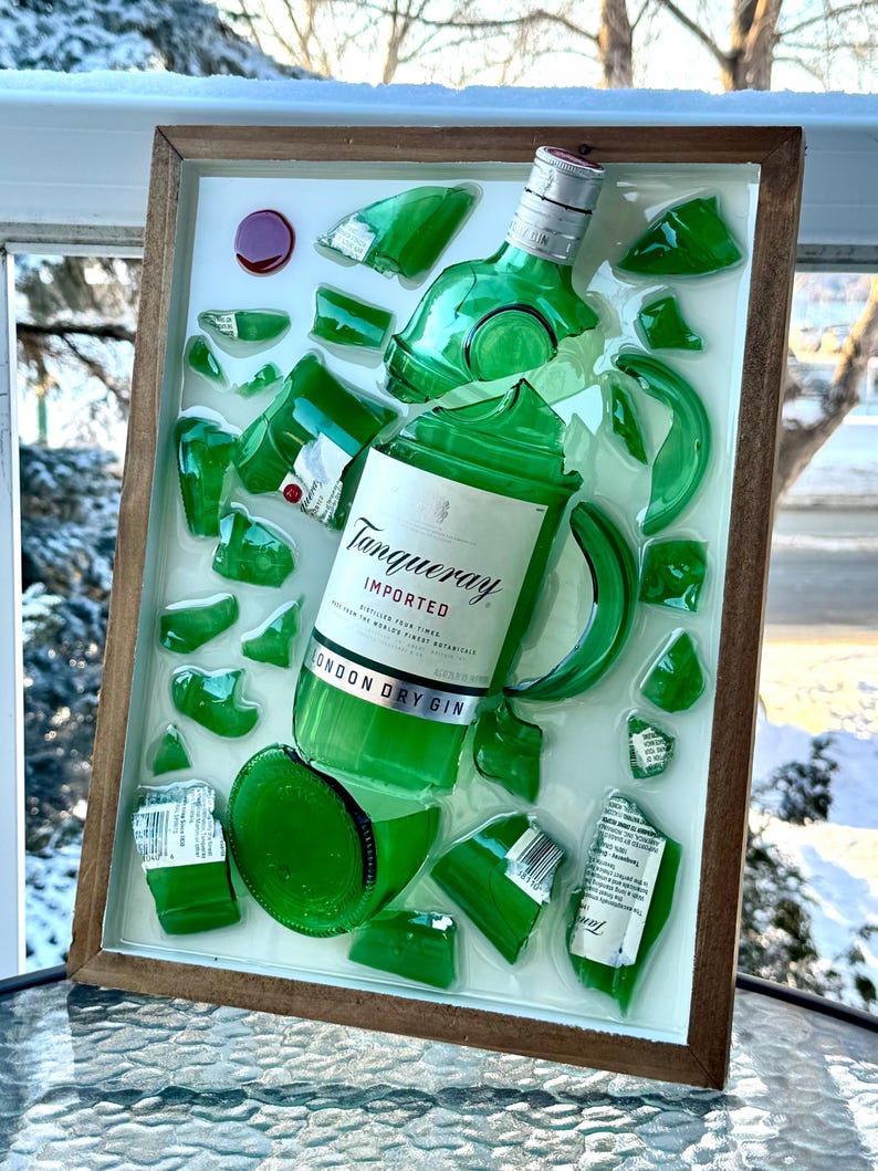 K&ouml;nnte beinhalten: Ein gerahmtes Kunstwerk mit einer zerbrochenen gr&uuml;nen Gin-Flasche, auf der das Etikett "Tanqueray London Dry Gin" zu sehen ist. Die Glasscherben sind auf wei&szlig;em Hintergrund in einem Holzrahmen angeordnet.