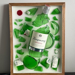K&ouml;nnte beinhalten: Ein gerahmtes Kunstwerk mit einer zerbrochenen gr&uuml;nen Gin-Flasche, deren Fragmente auf wei&szlig;em Hintergrund angeordnet sind. Das Etikett lautet "Tanqueray Imported London Dry Gin". Ein rotes Wachssiegel befindet sich oben links. Der Rahmen ist aus Holz.