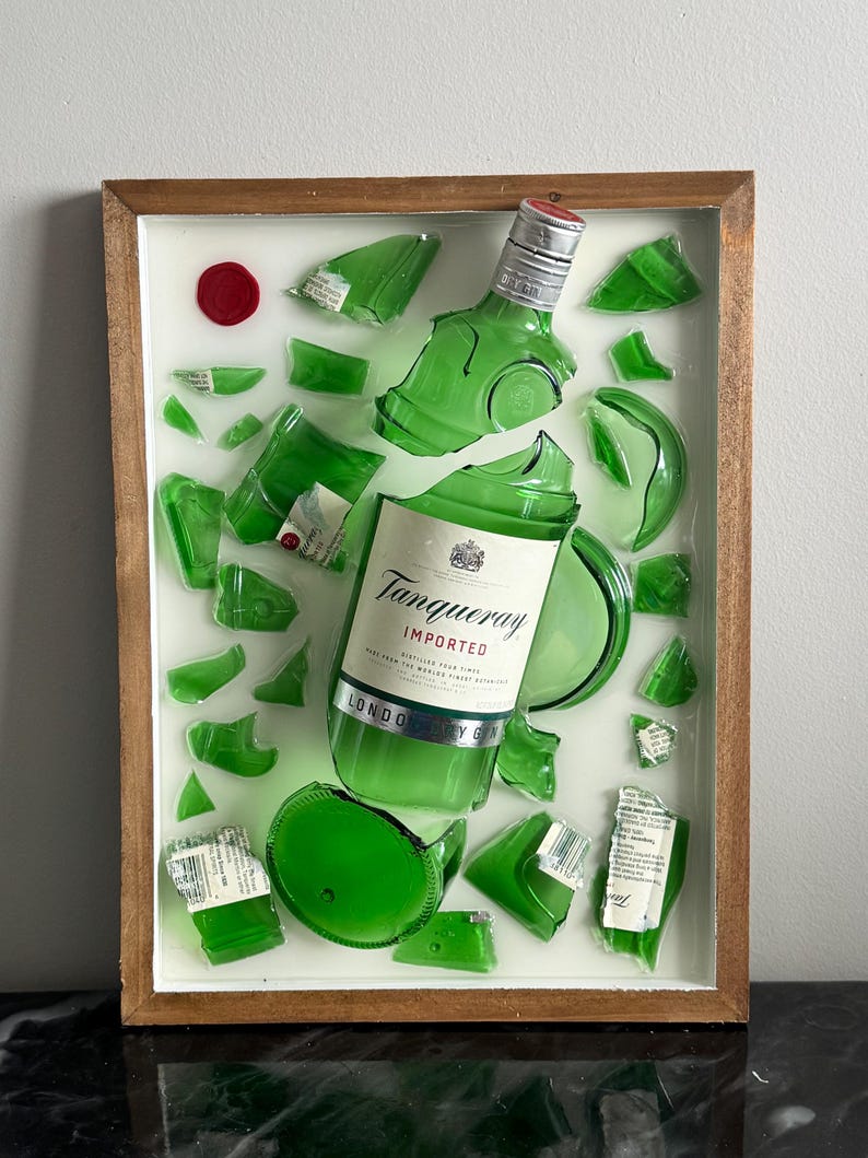 K&ouml;nnte beinhalten: Ein gerahmtes Kunstwerk mit einer zerbrochenen gr&uuml;nen Gin-Flasche, deren Fragmente auf wei&szlig;em Hintergrund angeordnet sind. Das Etikett lautet "Tanqueray Imported London Dry Gin". Ein rotes Wachssiegel befindet sich oben links. Der Rahmen ist aus Holz.