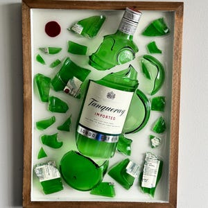K&ouml;nnte beinhalten: Gerahmtes Kunstwerk mit einer zerbrochenen gr&uuml;nen Glasflasche Tanqueray London Dry Gin. Die Flasche ist in mehrere Teile zerbrochen, das Etikett teilweise erhalten. Das Kunstwerk ist vor einem wei&szlig;en Hintergrund in einem Holzrahmen.
