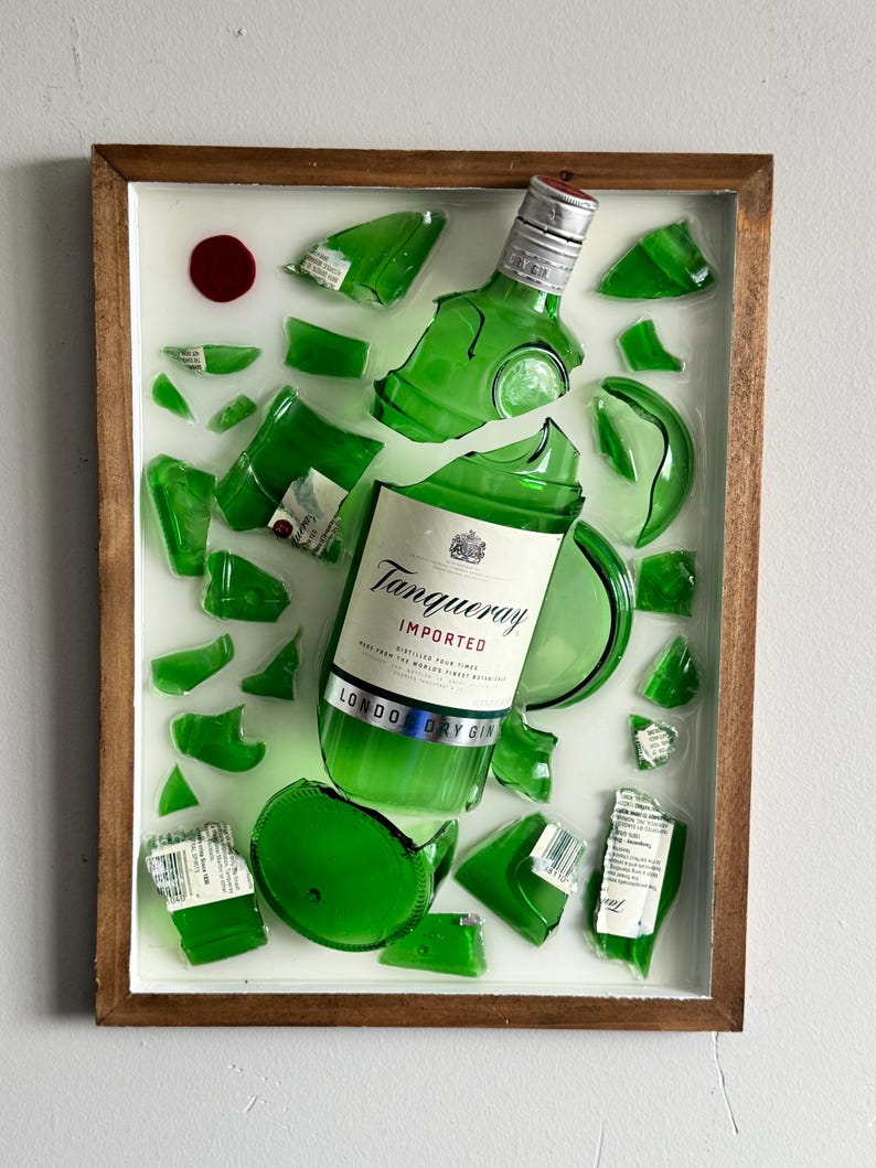K&ouml;nnte beinhalten: Gerahmtes Kunstwerk mit einer zerbrochenen gr&uuml;nen Glasflasche Tanqueray London Dry Gin. Die Flasche ist in mehrere Teile zerbrochen, das Etikett teilweise erhalten. Das Kunstwerk ist vor einem wei&szlig;en Hintergrund in einem Holzrahmen.