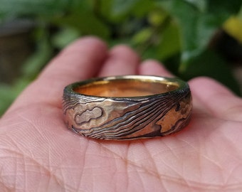 May include: A gold and silver patterned ring with a textured surface. The ring is worn on a person's finger.
