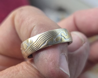 Puede incluir: Un anillo de metal con acabado bicolor, superficie plateada cepillada y zonas hundidas en color dorado pulido. La banda presenta un patrón similar a un mapa topográfico con líneas fluidas e irregulares y formas orgánicas.