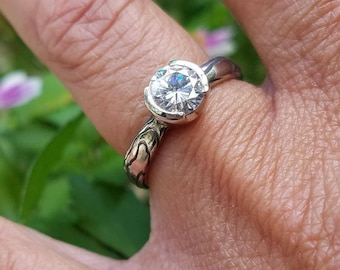 Puede incluir: Un anillo de tono plateado con una piedra redonda transparente y facetada en un engaste de bisel parcial. La banda presenta un patrón orgánico texturizado con líneas oscuras hundidas, contrastando con el acabado pulido del engaste.
