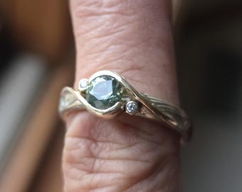 Puede incluir: Un anillo con una piedra central redonda y facetada de color verde oscuro montada en una banda de metal pulido. El diseño incluye dos piedras redondas más pequeñas y claras a los lados, con un engaste curvo que rodea la piedra central.