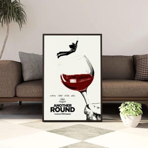 May include: A framed movie poster for Another Round featuring a large wine glass filled with red wine, a person jumping above the glass, and a hand holding the stem. The text reads Another Round, Thomas Vinterberg, and includes film credits.