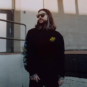 Puede incluir: Sudadera con capucha negra con un diseño cuadrado amarillo en el pecho y arte abstracto colorido en la manga izquierda. La persona de la imagen lleva gafas de sol y tiene el pelo largo.