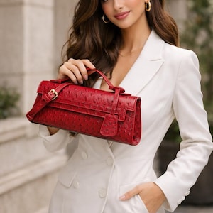 May include: A red leather handbag with a textured surface, a top handle, and a gold-coloured buckle. The bag has a rectangular shape and a flap closure. The person is wearing a white blazer.