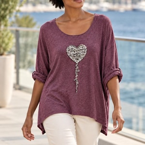 May include: A plum-coloured, long-sleeved top with a scoop neckline and a high-low hem. The front features a large, heart-shaped embellishment made of silver sequins and beads, with a dangling, beaded trail extending downwards from the heart's base.