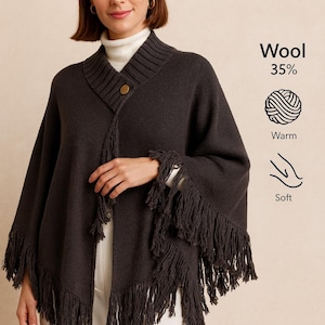Op de afbeelding: Een donkergrijze wollen poncho met franjes. De poncho heeft een knoopsluiting en is gelabeld als 35% wol, warm en zacht. Het model draagt er een witte coltrui onder.