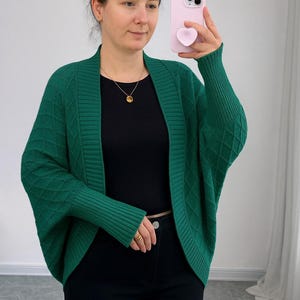 May include: A green cardigan with a diamond pattern. The cardigan has long sleeves and a wide collar. The person in the image is wearing a black top and black trousers. A gold necklace is visible.
