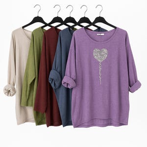 Könnte beinhalten: Fünf langärmlige Shirts in neutralen Farben hängen an schwarzen Kleiderbügeln. Die Shirts sind beige, olivgrün, weinrot, marineblau und lila. Das lila Shirt hat ein silbernes Herzdesign auf der Vorderseite.