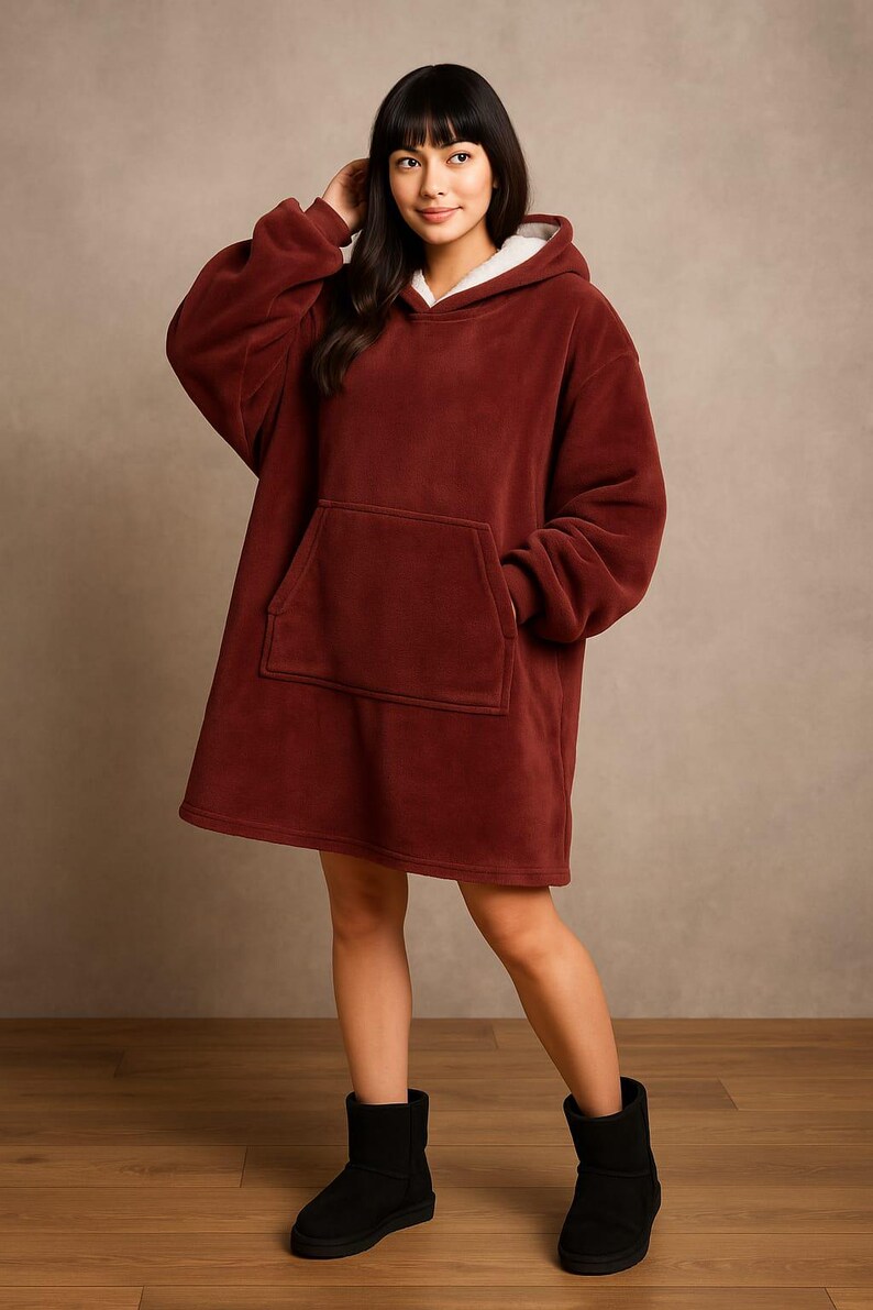 May include: A burgundy oversized hoodie with a kangaroo pocket and a white fleece-lined hood. The model is wearing black boots. The hoodie is made of a soft, plush material, perfect for lounging.