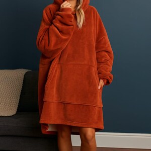May include: A rust-colored oversized hooded blanket, featuring a large front pocket and a cozy, plush texture. The hooded blanket is designed for comfort and warmth, perfect for lounging. The model is wearing the blanket indoors.
