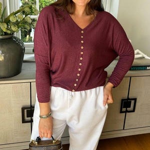 May include: A burgundy long-sleeved top with a V-neck and button-down front, paired with white drawstring trousers. A small, patterned handbag is held in the hand. The outfit is casual and comfortable.