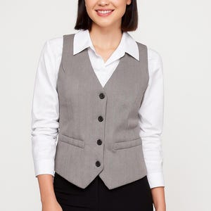 Puede incluir: Un chaleco gris con estampado de espiga con cuatro botones negros, usado sobre una camisa blanca con cuello y pantalones negros. El chaleco tiene dos bolsillos delanteros. El atuendo es informal de negocios.
