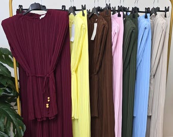 May include: A clothing rack displays a variety of pleated two-piece outfits in various colors. The outfits include a burgundy set with a matching belt, a yellow set, a brown set, a pink set, a green set, a blue set, and a beige set.