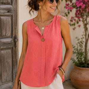 Puede incluir: Una blusa sin mangas de color coral con escote en V y un solo botón marrón de cierre en el centro. La prenda tiene un corte holgado con un dobladillo redondeado y una textura de tela visible, combinada con pantalones de color beige claro.