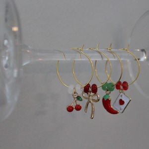 May include: Gold wine charms with decorative charms, including cherries, a bow, a chilli pepper, and a playing card. The charms are suspended on a clear glass stem.
